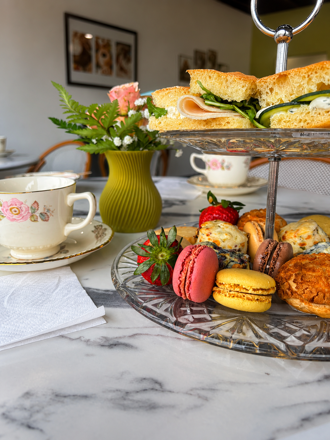 An elegant afternoon tea spread is beautifully arranged on a white marble table inside a bright, cozy café. A two-tier clear glass serving stand takes center stage. On the top tier are thick, golden focaccia sandwich halves layered with sliced deli meat, fresh arugula, creamy white cheese, and thin cucumber ribbons, showcasing the airy texture of the bread. The lower tier features an assortment of colorful French macarons in pink, yellow, and chocolate brown, fresh ripe strawberries with green tops, golden 