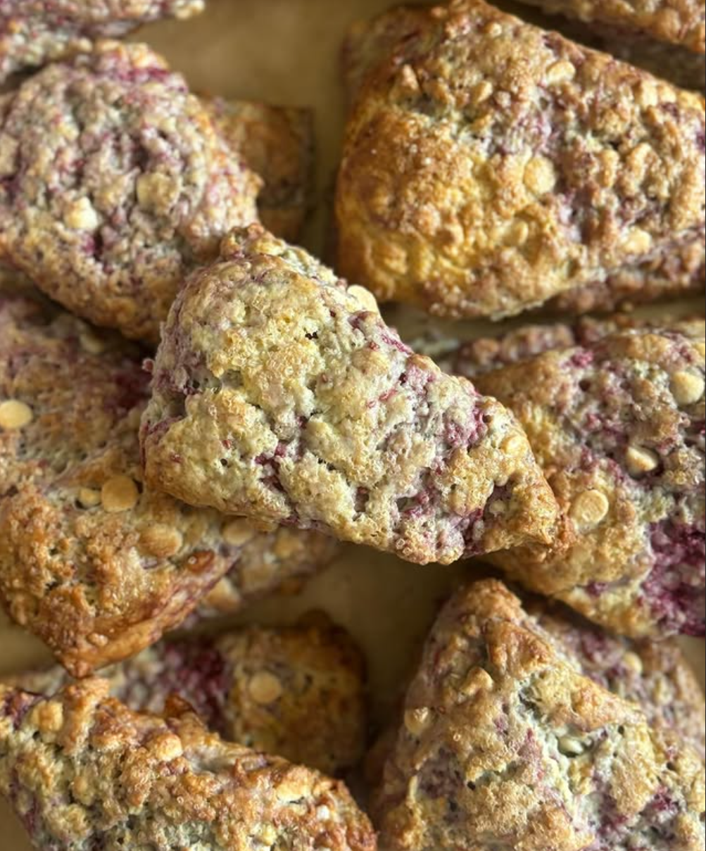A close-up view of freshly baked artisan berry scones arranged on a parchment-lined baking tray. The triangular scones are golden brown with crisp, rustic edges and generously topped with a buttery crumb streusel that adds a crunchy texture. Bursts of vibrant red berries are visible throughout the flaky interior, creating a beautiful contrast against the light, tender crumb. White chocolate chips are scattered across the tops, adding a touch of sweetness and visual appeal.

The scones have an uneven, handcr