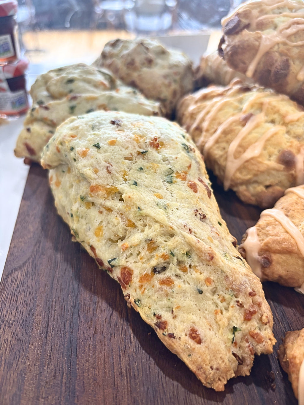 A close-up photograph of a freshly baked savory artisan scone resting on a dark wooden serving board inside a bakery display. The triangular scone has a lightly golden crust with a soft, fluffy interior visible at the cut edge. It is generously filled with melted cheddar cheese, herbs, and savory bacon bits, creating a speckled appearance throughout the crumb. The texture looks tender and flaky, with a crisp exterior and moist center.

In the softly blurred background are additional freshly baked scones, in