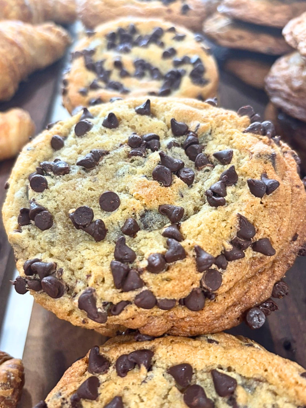 A close-up photograph of a freshly baked gourmet chocolate chip cookie displayed on a dark wooden bakery tray. The oversized cookie is golden brown with crisp, lightly caramelized edges and a soft, thick center generously covered with semi-sweet chocolate chips. Additional chocolate chips are baked throughout the dough, creating a rich, indulgent appearance with pockets of melted chocolate in every bite. The cookie's cracked surface and slightly domed shape highlight its chewy interior and handcrafted, bake
