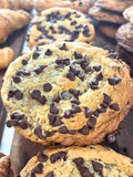 A close-up photograph of a freshly baked gourmet chocolate chip cookie displayed on a dark wooden bakery tray. The oversized cookie is golden brown with crisp, lightly caramelized edges and a soft, thick center generously covered with semi-sweet chocolate chips. Additional chocolate chips are baked throughout the dough, creating a rich, indulgent appearance with pockets of melted chocolate in every bite. The cookie's cracked surface and slightly domed shape highlight its chewy interior and handcrafted, bake